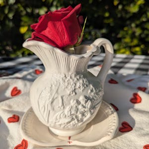 May include: White ceramic pitcher with a raised design of a couple and a tree. A single red rose is placed in the pitcher. The pitcher is sitting on a white saucer with a heart-shaped design.