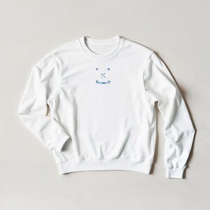 K&ouml;nnte beinhalten: Wei&szlig;es Sweatshirt mit Rundhalsausschnitt und einer kleinen blauen Grafik auf der Brust. Die Grafik zeigt zwei gekreuzte Pfeile, zwei V&ouml;gel und ein Banner. Das Sweatshirt hat lange &Auml;rmel und gerippte B&uuml;ndchen und Saum.