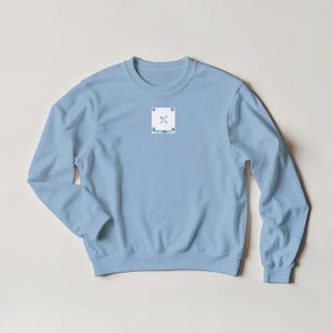 K&ouml;nnte beinhalten: Hellblaues Sweatshirt mit einem kleinen quadratischen Aufn&auml;her auf der Brust. Das Design zeigt gekreuzte Pfeile und stilisierte Bl&auml;tter in Blau und Wei&szlig;. Das Sweatshirt hat lange &Auml;rmel und einen Rundhalsausschnitt.