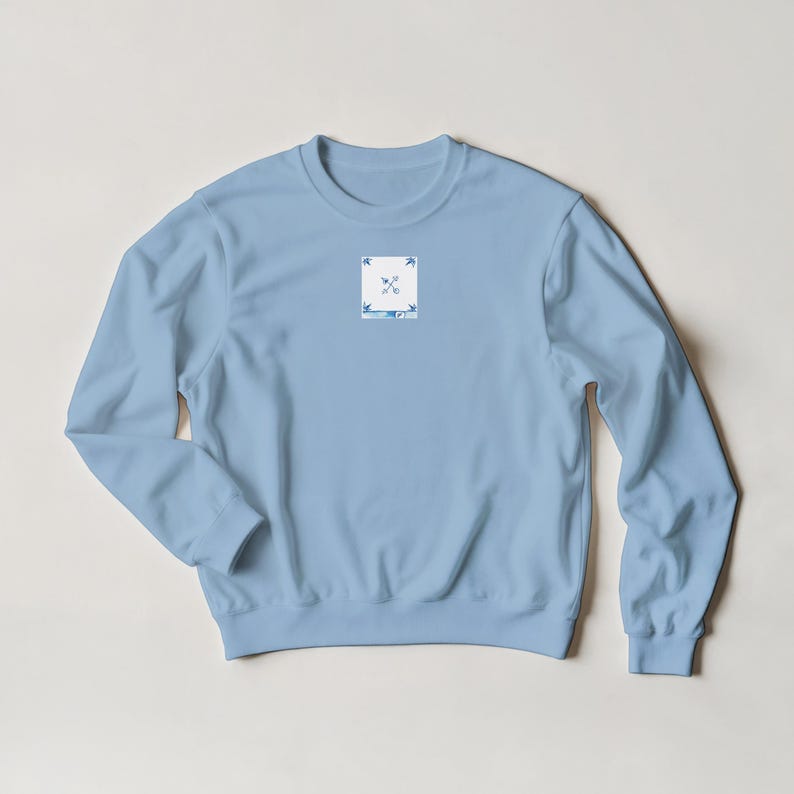 K&ouml;nnte beinhalten: Hellblaues Sweatshirt mit einem kleinen quadratischen Aufn&auml;her auf der Brust. Das Design zeigt gekreuzte Pfeile und stilisierte Bl&auml;tter in Blau und Wei&szlig;. Das Sweatshirt hat lange &Auml;rmel und einen Rundhalsausschnitt.
