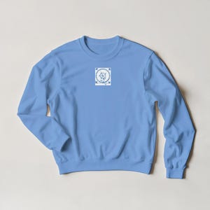 K&ouml;nnte beinhalten: Hellblaues Sweatshirt mit Rundhalsausschnitt und langen &Auml;rmeln. Das Sweatshirt hat eine kleine, quadratische Grafik auf der Brust mit einem blau-wei&szlig;en Blumendesign. Die Grafik enth&auml;lt den Schriftzug "love" in Schreibschrift.