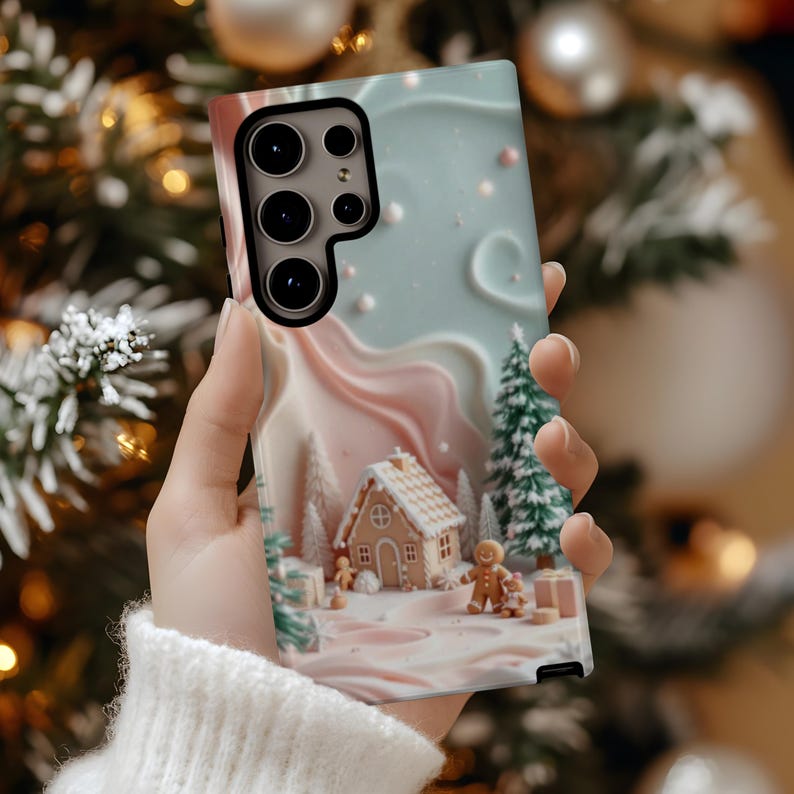 May include: A phone case featuring a winter gingerbread scene with a house, gingerbread people, and Christmas trees. The case has a light blue and pink color scheme with a 3D effect. The phone case is held in a person's hand.