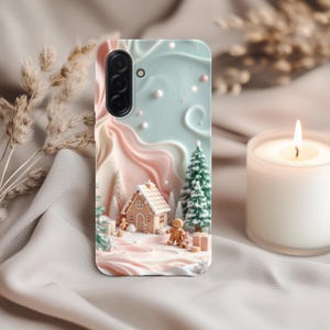 May include: A phone case featuring a winter scene with a gingerbread house, gingerbread man, and Christmas trees. The case has a light blue and pink background with a textured design. A lit candle is in the background.