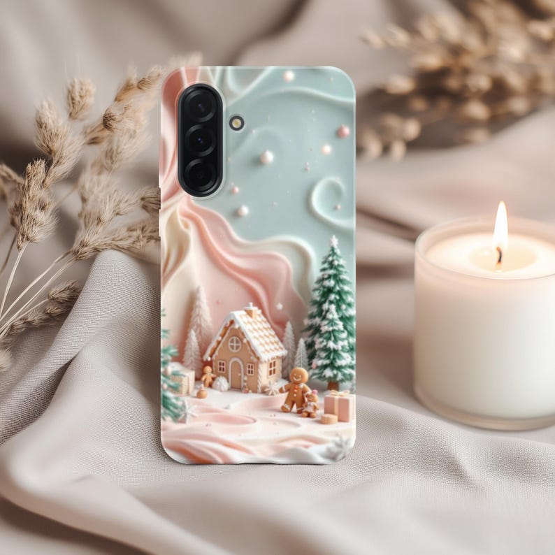 May include: A phone case featuring a winter scene with a gingerbread house, gingerbread man, and Christmas trees. The case has a light blue and pink background with a textured design. A lit candle is in the background.
