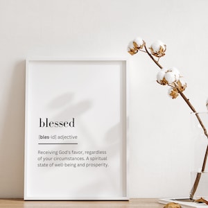 Op de afbeelding: Een witte ingelijste print met het woord "blessed" in zwarte tekst, gevolgd door een definitie in kleiner lettertype. De definitie luidt: "[bles-id] bijvoeglijk naamwoord Gods gunst ontvangen, ongeacht je omstandigheden. Een spirituele toestand van welzijn en voorspoed."