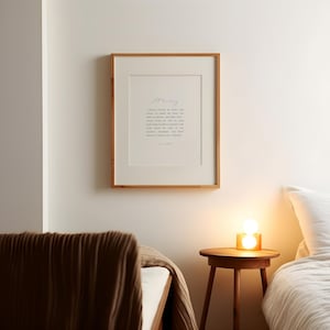 May include: A framed print with a light wood frame hangs on a white wall. The print features a poem in black text on a white background. A small wooden side table with a lamp sits below the print. A bed with a brown blanket is in the foreground.