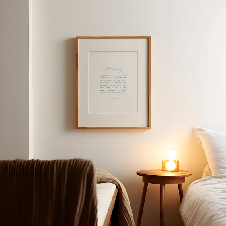May include: A framed print with a light wood frame hangs on a white wall. The print features a poem in black text on a white background. A small wooden side table with a lamp sits below the print. A bed with a brown blanket is in the foreground.