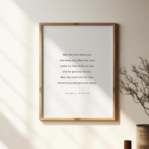 Könnte beinhalten: Ein gerahmter Druck mit einem hellen Holzrahmen. Der Druck hat einen weißen Hintergrund mit schwarzem Text, der lautet: "May the Lord bless you...Numbers 6:24-26". Der Druck wird an einer weißen Wand angezeigt.