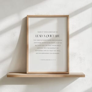 May include: A framed print with a light wood frame and a white background. The print features black text that reads "MAKE IT YOUR AMBITION TO LEAD A QUIET LIFE" and a quote from 1 Thessalonians 4:11-12. The framed print is displayed on a wooden shelf.