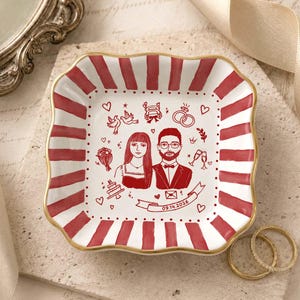 Puede incluir: Un plato de cer&aacute;mica cuadrado con bordes festoneados y un borde pintado en oro. El plato presenta rayas rojas y blancas a lo largo del borde y una ilustraci&oacute;n central de l&iacute;nea roja de una pareja, iconos de boda y una pancarta con la fecha 09.14.2026.