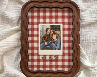 May include: A rectangular wooden frame with a wavy edge containing a red and white gingham background. Centered is a polaroid-style illustration of a couple sitting together, with the text "Theo & Sadie" and "07.24.2026" printed below the image.