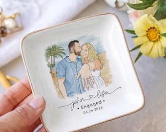 May include: A square white ceramic trinket dish with a gold-toned rim featuring a watercolor illustration of a couple on a beach. The woman displays an engagement ring. Text reads: john lisa • Engaged • 06.24.2026 in black script and sans-serif font.