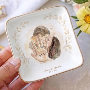 May include: A square white ceramic trinket dish with a gold-toned rim. The center features a watercolor illustration of a couple kissing, overlaid with a hand wearing a ring and the text I SAID yes!. Below, the names James & Amelia and 04.20.2026.