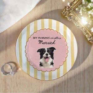 May include: A round ceramic plate featuring vertical yellow and white stripes. The center has a pink scalloped circle with a black and white border collie illustration and the text "MY HUMANS are getting Married" in black lettering.
