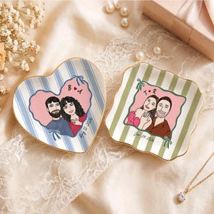 May include: Two ceramic trinket dishes with gold-painted edges. One heart-shaped dish features blue stripes and a couple illustration with the text B A and Aug 12, 2026. The other square dish has green stripes and a couple illustration with T R and Jan 24, 2026.