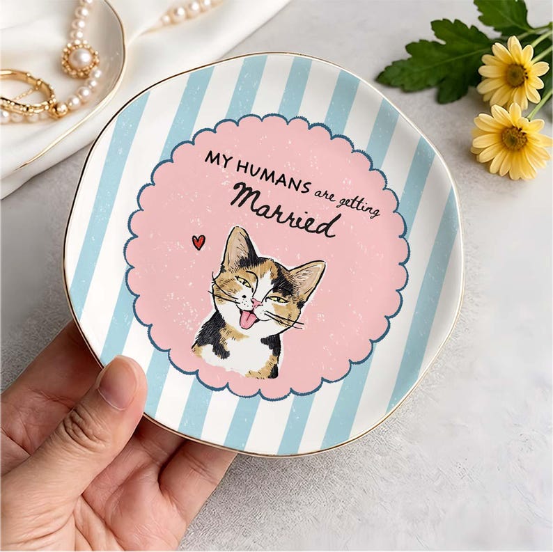 May include: A round ceramic trinket dish featuring light blue and white vertical stripes. The center has a pink scalloped circle with a calico cat illustration, a small red heart, and the text "MY HUMANS are getting Married" in black.