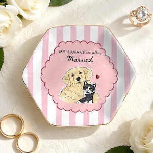May include: A hexagonal ceramic ring dish featuring a pink and white striped pattern with a scalloped central frame. The text reads MY HUMANS are getting Married above an illustration of a golden retriever and a black and white cat with a small red heart.