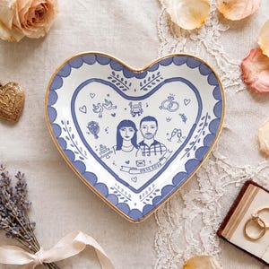 Puede incluir: Un plato de cer&aacute;mica en forma de coraz&oacute;n con borde dorado y cenefa festoneada azul. El centro presenta una ilustraci&oacute;n lineal de una pareja, iconos de boda y una cinta con la fecha 09.14.2026 impresa en tinta azul oscuro.