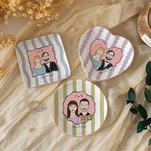 Pu&ograve; includere: Tre piattini in ceramica con illustrazioni personalizzate di coppie. Un piattino quadrato a righe blu, uno a forma di cuore a righe viola e uno rotondo a righe verdi. Ogni piattino mostra un ritratto, le iniziali T R e July 24 2026.
