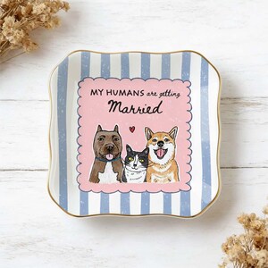 May include: A square ceramic trinket dish with scalloped edges and a gold-toned rim. The background features light blue and white vertical stripes. A central pink scalloped square displays the text MY HUMANS are getting Married with illustrations of a brown dog, a black and white cat, and a tan dog.