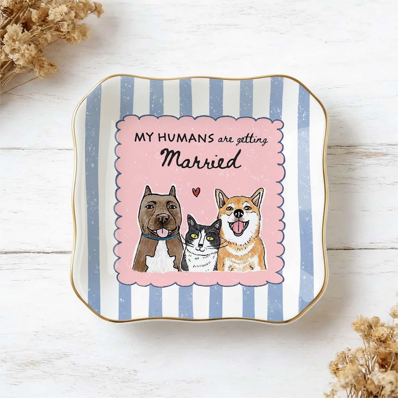 May include: A square ceramic trinket dish with scalloped edges and a gold-toned rim. The background features light blue and white vertical stripes. A central pink scalloped square displays the text MY HUMANS are getting Married with illustrations of a brown dog, a black and white cat, and a tan dog.