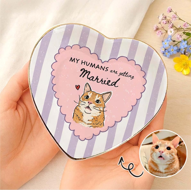 May include: A heart-shaped ceramic trinket dish featuring a light purple and white vertical striped pattern. The center displays a pink scalloped heart with a cartoon orange tabby cat illustration and the text MY HUMANS are getting Married.