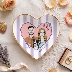 May include: A heart-shaped ceramic trinket dish featuring a gold rim and a light purple striped background. The center displays a custom illustration of a couple and a black and white dog inside a pink heart, with the letters B and A and the date Aug 12 2026.