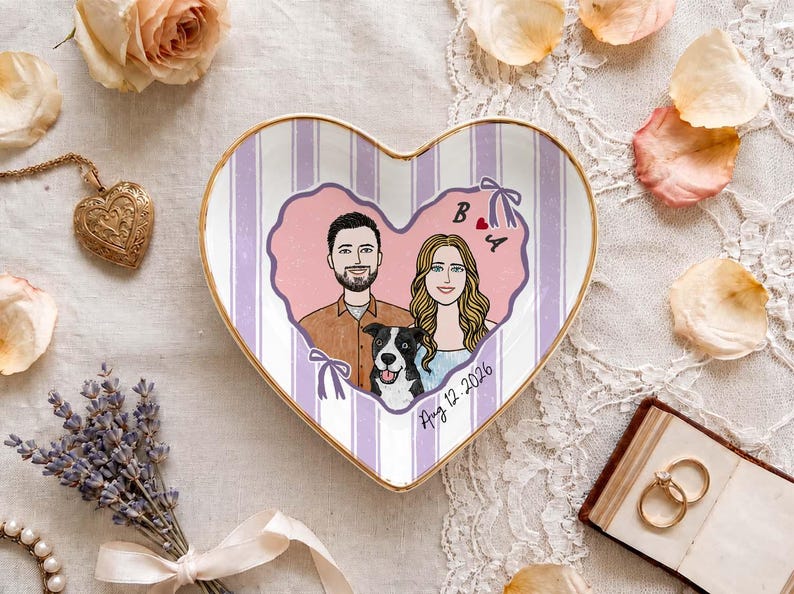 May include: A heart-shaped ceramic trinket dish featuring a gold rim and a light purple striped background. The center displays a custom illustration of a couple and a black and white dog inside a pink heart, with the letters B and A and the date Aug 12 2026.