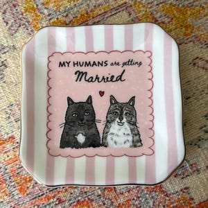 May include: A square ceramic trinket dish with rounded corners, featuring a light pink and white vertical striped border. The center displays two illustrated cats, one solid grey and one tabby, with the text "MY HUMANS are getting Married" and a small red heart.