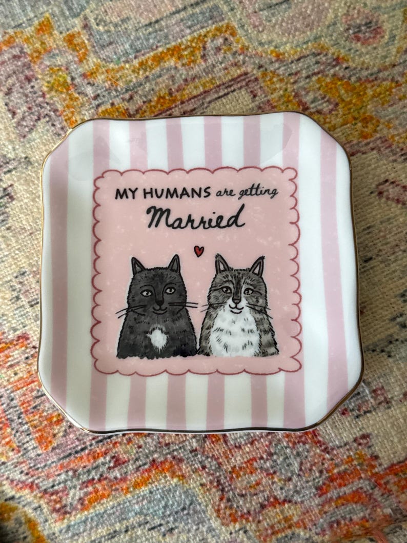 May include: A square ceramic trinket dish with rounded corners, featuring a light pink and white vertical striped border. The center displays two illustrated cats, one solid grey and one tabby, with the text "MY HUMANS are getting Married" and a small red heart.