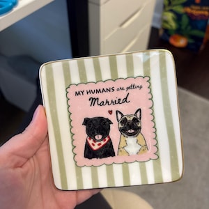 May include: A square ceramic trinket dish with a gold-trimmed edge, featuring a light green and white vertical striped background. The center displays a pink scalloped frame with two illustrated dogs and the text, MY HUMANS are getting Married.