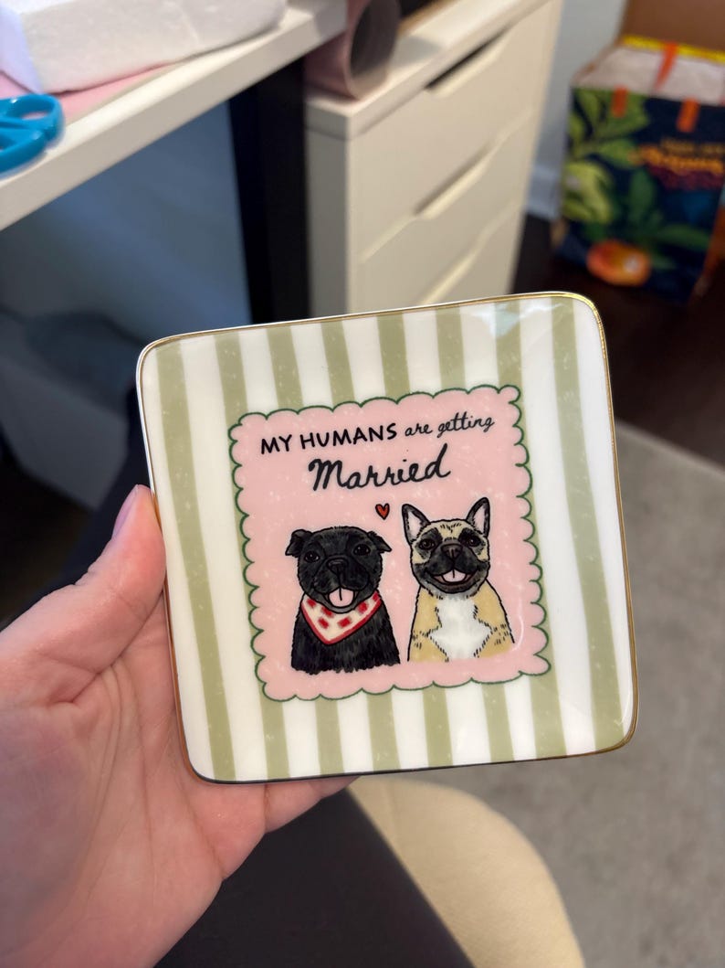 May include: A square ceramic trinket dish with a gold-trimmed edge, featuring a light green and white vertical striped background. The center displays a pink scalloped frame with two illustrated dogs and the text, MY HUMANS are getting Married.