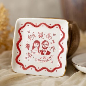 Puede incluir: Un plato cuadrado de cer&aacute;mica blanca con borde dorado y una l&iacute;nea roja ondulada. El centro presenta ilustraciones en l&iacute;nea roja de una pareja, anillos de boda, palomas, un coche, un pastel y una cinta con la fecha 11.26.2026 impresa en &eacute;l.