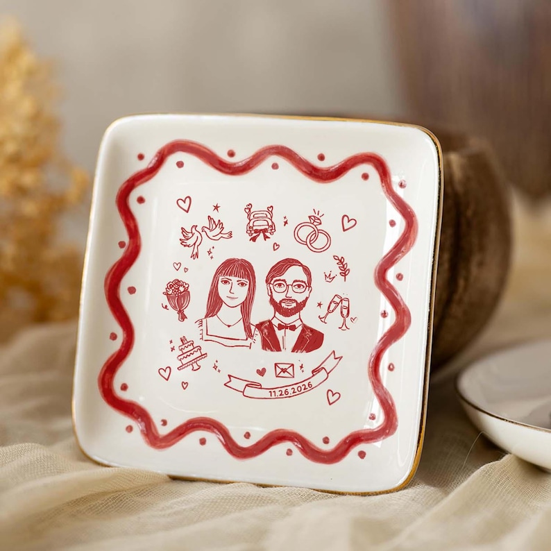 Puede incluir: Un plato cuadrado de cer&aacute;mica blanca con borde dorado y una l&iacute;nea roja ondulada. El centro presenta ilustraciones en l&iacute;nea roja de una pareja, anillos de boda, palomas, un coche, un pastel y una cinta con la fecha 11.26.2026 impresa en &eacute;l.