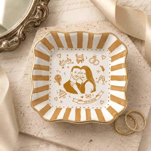 Puede incluir: Un plato decorativo cuadrado de cer&aacute;mica con bordes festoneados y rayas verticales pintadas en dorado. El centro muestra una ilustraci&oacute;n de l&iacute;nea dorada de una pareja rodeada de iconos de boda y una cinta con la fecha 09.14.2026 impresa.