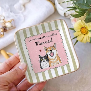 May include: A square ceramic trinket dish with a gold-toned rim, featuring a light green and white striped border. The center displays a pink scalloped square with a black and white cat, a tan dog, and the text "MY HUMANS are getting Married".