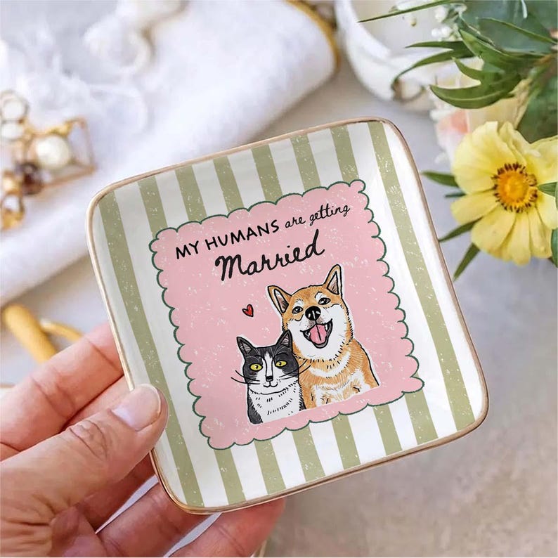 May include: A square ceramic trinket dish with a gold-toned rim, featuring a light green and white striped border. The center displays a pink scalloped square with a black and white cat, a tan dog, and the text "MY HUMANS are getting Married".