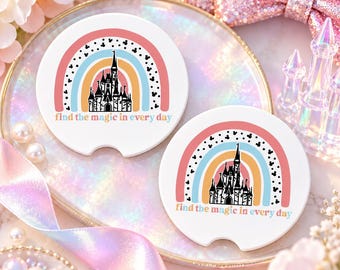 Puede incluir: Dos posavasos redondos blancos para coche con un gráfico de arcoíris colorido, una silueta de castillo negra y pequeños iconos de orejas de ratón negros. El texto "find the magic in every day" está impreso en minúsculas en la parte inferior.