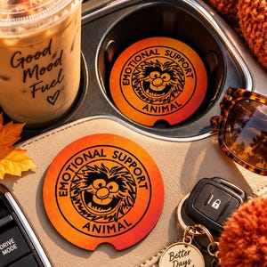 May include: Two circular car cup holder coasters featuring a gradient orange and red background. Each coaster displays a black graphic of a furry character with the text "EMOTIONAL SUPPORT ANIMAL" printed in a circular black font around the character.