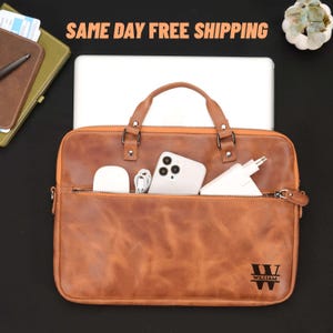 Op de afbeelding: Een bruine leren laptoptas met een ritsvak met een witte telefoon, oplader en draadloze oordopjes. De tas heeft een handvat en de naam "WILLIAM" staat rechtsonder gedrukt.