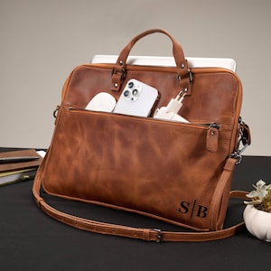 Könnte beinhalten: Eine braune Laptoptasche aus Leder mit einer vorderen Reißverschlusstasche, zwei Tragegriffen und einem verstellbaren Schultergurt. Die Tasche ist mit den Initialen S|B unten rechts personalisiert und enthält ein Smartphone sowie eine weiße Maus.