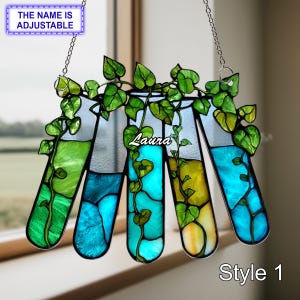 Op de afbeelding: Gebrandschilderd glaskunstwerk met vijf reageerbuisvormen, elk gevuld met gekleurde vloeistof en versierd met groene wijnstokken. De naam "Laura" is zichtbaar. De tekst "THE NAME IS ADJUSTABLE" en "Style 1" zijn ook zichtbaar.