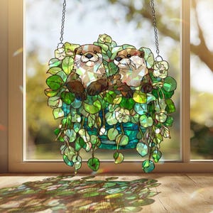 Puede incluir: Adorno de ventana de vidrieras con dos nutrias en una cesta colgante de follaje verde y blanco. Las nutrias tienen pelaje marrón. La pieza cuelga de una cadena.