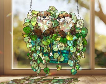 May include: Stained glass window hanging featuring two otters nestled in a hanging basket of green and white foliage. The otters have brown fur and are surrounded by leaves and flowers. The piece hangs from a chain.