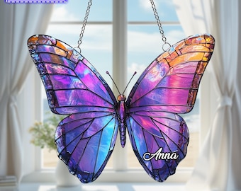 May include: A stained glass butterfly suncatcher featuring iridescent purple, blue, and orange wings with black lead lines. The name Anna is printed on the lower right wing. A metal chain is attached for hanging. Text reads: THE NAME IS ADJUSTABLE.