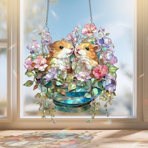 Può includere: Decorazione per finestra in vetro colorato raffigurante due criceti in un vaso pieno di fiori. Il vaso è blu e verde, con fiori rosa, viola e bianchi. I criceti sono marroni e bianchi, appesi a una catena.