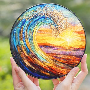 May include: A round, stained glass art piece depicting a breaking ocean wave with a setting sun. The artwork features vibrant blues, oranges, and yellows, with a black metal frame. The piece is held up by two hands.