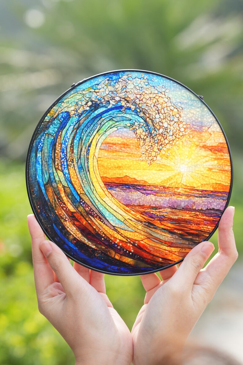 May include: A round, stained glass art piece depicting a breaking ocean wave with a setting sun. The artwork features vibrant blues, oranges, and yellows, with a black metal frame. The piece is held up by two hands.