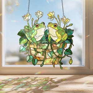 May include: Stained glass window decoration featuring two green frogs in a hanging basket with white and yellow flowers and green leaves. The artwork is suspended by chains and is set against a window.