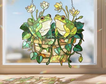 May include: Stained glass window decoration depicting two green frogs in a hanging basket with white and yellow flowers and green leaves. The artwork is suspended by chains and set against a window.